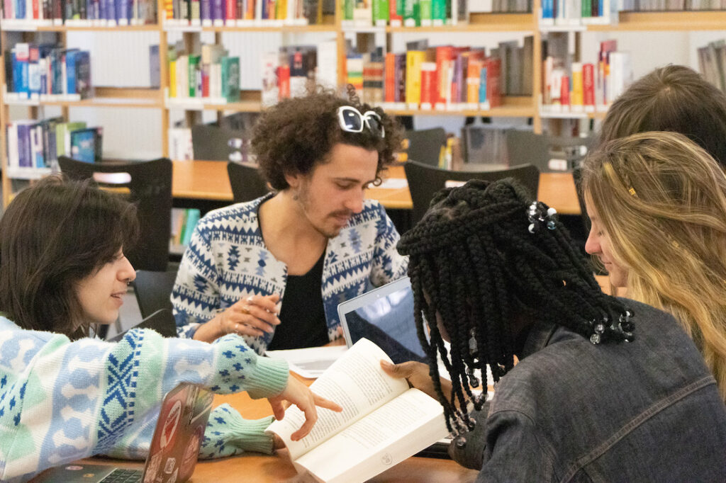 Un groupe de 5 travaillent dans la bibliothèque. Une des élèves montre du doigts une phrase sur un livre ouvert