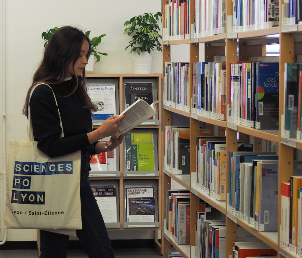 Une élève lit un livre debout à coté d'une étagère de la bibliothèque, un tote bag sciences po sur l'épaule